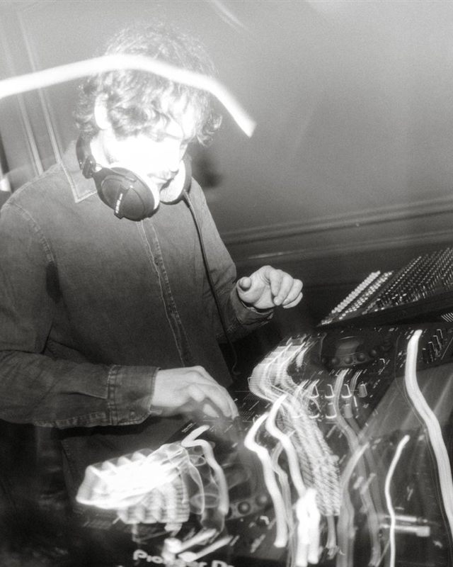 Diego Ramirez DJing, headphones on, working the mixer under a long exposure of light trails.